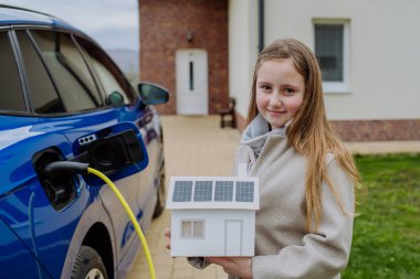 Güneş panelleri olan kağıt bir evin modelini tutan küçük bir kız elektrikli arabalarını şarj etmek için bekliyor..