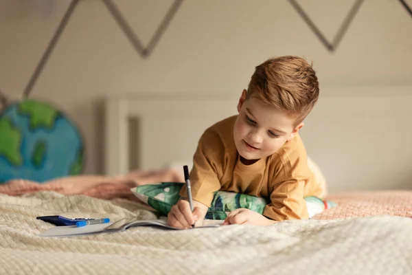 Küçük Çocuk Yatakta Uzanıyor Ödevini Yapıyor — Stok fotoğraf