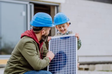 Close-up of father and his little son holding solar panel, in front of their unfinished house. clipart