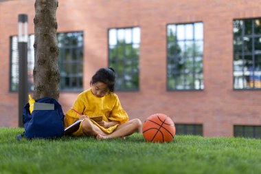 Little asian girl sitting in public park and writing some notes. Summer time.