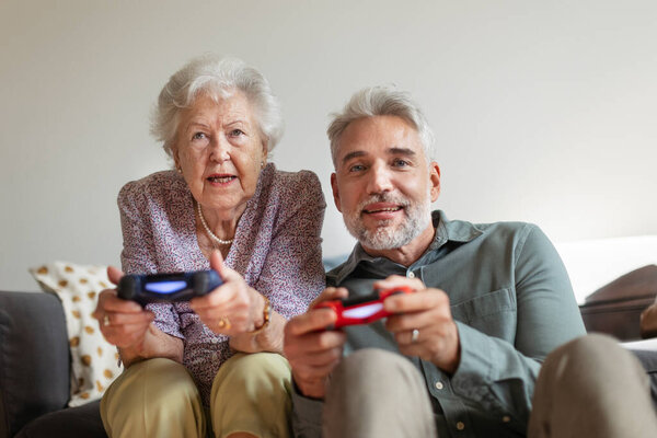 Mature man playing video games with his senior mother, having fun. Adult son enjoys spending time with his elderly mother.