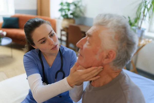 医師は首リンパ節を調べる 老人ホームで介護する若い看護師 — ストック写真