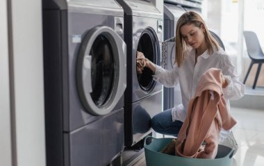 Young woman loading washing machine in a public laundry. clipart