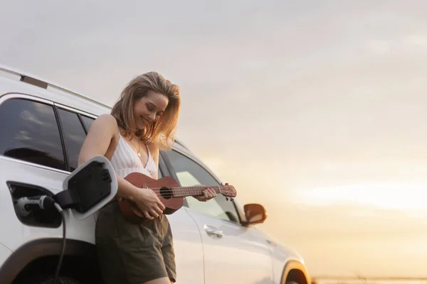 一个年轻女子靠在车上 玩四弦琴 享受夏日时光的画像 — 图库照片