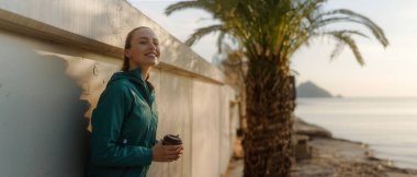 Sabah antrenmanından sonra plajda dinlenen genç bir fitness kadını. Güzel, sportif bir kadın, deniz kenarında güneşin doğuşunu ve bir fincan kahvenin tadını çıkarıyor..