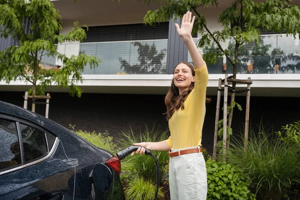 stock image Close up of beautiful woman waving, greeting someone while charging her electric car in front of her house. Pluging in charger in charging port.