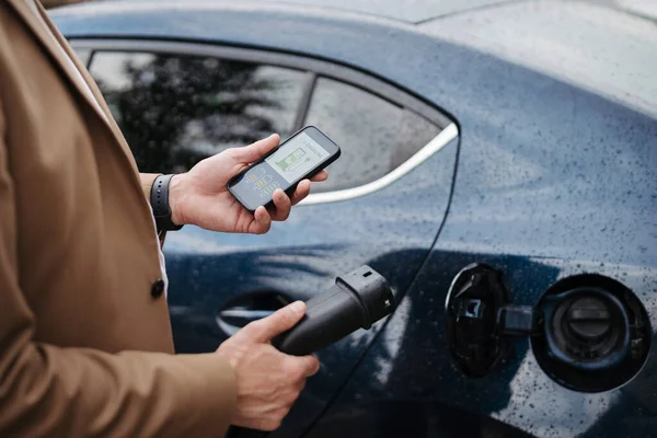 電気自動車充電アプリを使用してビジネスマンのクローズアップ スマートフォンから彼の電気自動車の充電をチェック 充電ステーションを見つける 電気使用量を監視するための充電アプリ — ストック写真
