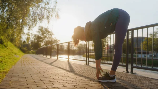 年轻的女赛跑选手在大清早跑城市之前伸展着胳膊和腿 穿着运动服的健身女孩准备晚上的运动 户外运动的概念 晨曦中的轮廓 — 图库照片