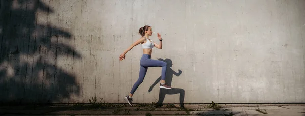 スポーツウェアを着た美しい女性選手が市内で運動している コンクリートの壁の影の前で実行しています コピースペース付きバナー — ストック写真