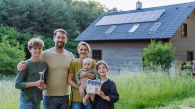 Mutlu bir aile rüzgar türbini ve fotovoltaikli kağıt ev modeli tutuyor. Evlerinin yanında güneş panelleri var. Alternatif enerji, tasarruf kaynakları ve sürdürülebilir yaşam tarzı kavramı.