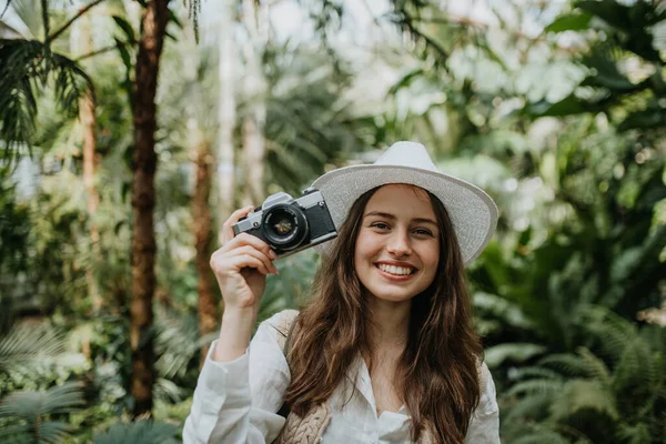 植物園で帽子とバックパックを持つ若い女性写真家の肖像画 温室内の植物学者が写真を撮る — ストック写真