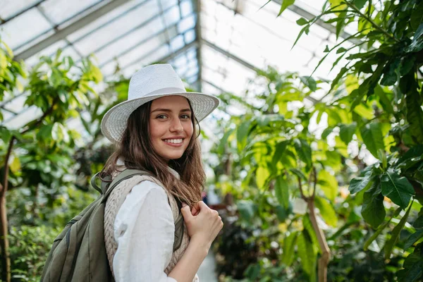 一个带着帽子和背包的年轻女子在植物园中的画像 温室植物学家 — 图库照片