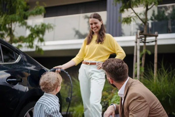 路上で電気自動車を充電する女性の肖像画 夫と息子が彼女を見ている間 電気自動車を充電するのを待っているフロントハウスの幸せな家族 — ストック写真