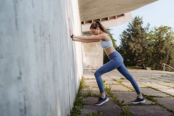 アウトドアを刺激するスポーツウェアの若いスポーティーな女性のサイドビュー 市内のコンクリート壁に向かって伸びるフィットネス女性 健康的なライフスタイルのコンセプト ストリートワークアウト — ストック写真