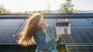 Çatıda güneş panelleri olan genç bir kadının portresi, fotovoltaikli ev modelini tutuyor..