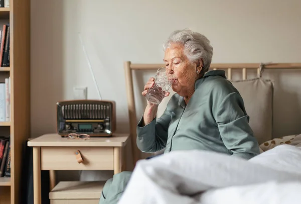 老年妇女穿着睡衣坐在床上喝水 — 图库照片
