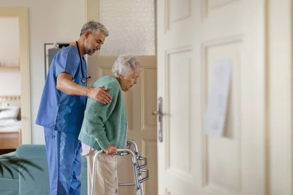 Caregiver helping senior woman to walk in her home. Thoughful male nurse taking care of eldery patient with walker.