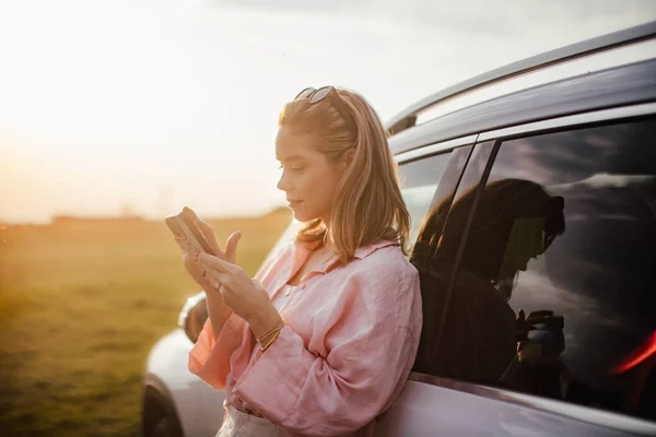 Молодая Женщина Прокручивает Телефон Опираясь Свою Машину Природе — стоковое фото