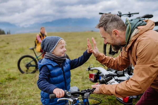 Young family with little children preparing for bicycle ride in nature, putting off bicycles from car racks. Father and son high five, looking forward to ride together. Concept of healthy lifestyle