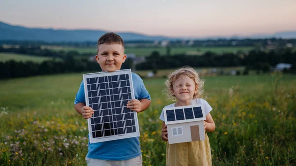 Young boy holding solar panel and his sister holding model of house with installed solar panels. Alternative energy, saving resources and sustainable lifestyle concept. Siblings standing in the middle