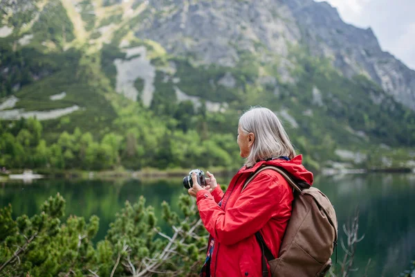 年富力强的老年女子在秋山远足的形象 在老年友好的小径上 带背包的资深游客用数码相机拍照 — 图库照片