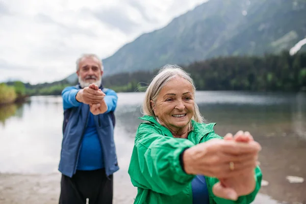 Senior couple doing outdoor yoga, tai chi, pilates by the lake in the autumn. Elderly husband and wife spending active vacation in the mountains, enjoying combination of physical activity and
