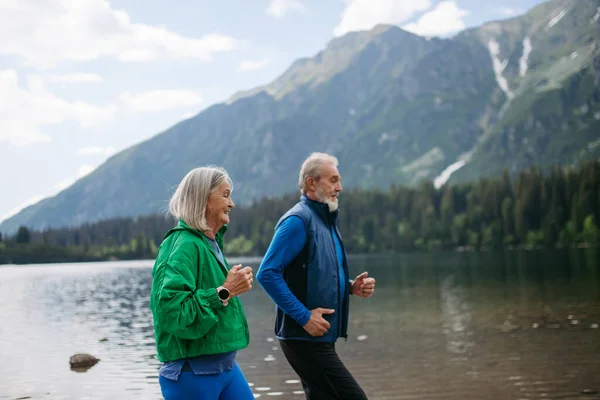 Senior couple running by the lake in the autumn. Elderly husband and wife spending active vacation in the mountains, enjoying combination of physical activity and relaxation.