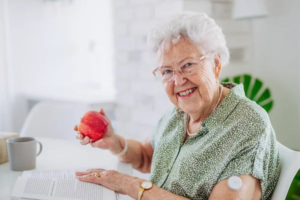 糖尿病老年患者使用连续血糖监测器检查家里的血糖水平 老年妇女吃苹果是为了帮助把血糖提高到正常水平 使老年妇女生活的Cgm装置 — 图库照片