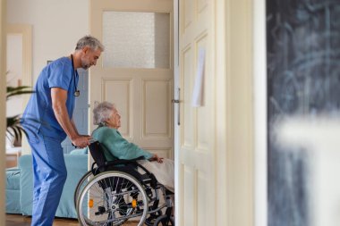 Bakıcı yaşlı kadını tekerlekli sandalyeye itiyor, hastanın evine taşınmasına yardım ediyor. Düşünceli erkek hemşire tekerlekli sandalyedeki yaşlı hastayla ilgileniyor..