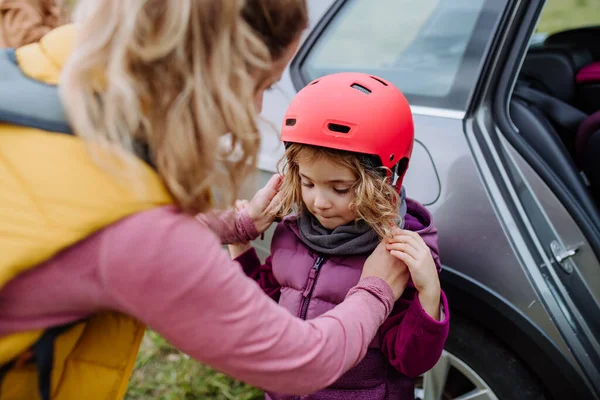 Mother putting on protective helmet on head of her daughter. Young family with little children preparing for bicycle ride in nature. Concept of healthy lifestyle and moving activity.