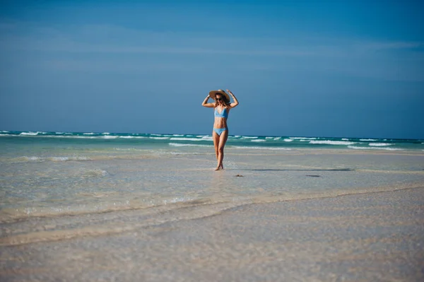 一个穿着比基尼和草帽站在海滩上的美丽瘦小女人的画像 身着蓝色泳衣 戴着太阳镜 享受沙滩和水晶般的Mnemba海滩的女子全身照 — 图库照片