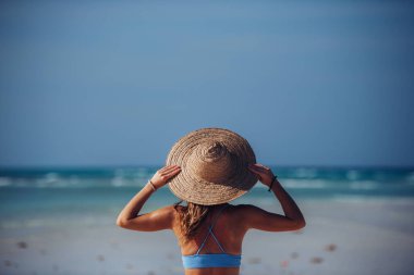Sahilde bikini ve hasır şapkalı güzel, ince bir kadının dikiz manzarası. Mavi mayo giymiş, kumlu plajın ve Zanzibar 'daki Mnemba plajının kristal denizinin tadını çıkaran bir kadının belden yukarı pozu.