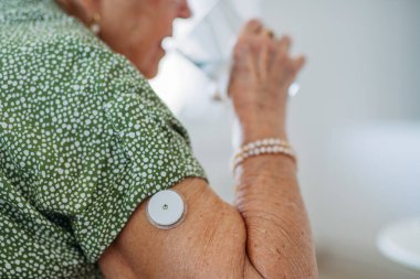 Diyabetik kıdemli hasta evdeki kan şekerini ölçmek için sürekli glikoz monitörü kullanıyor. Kıdemli kadın diyabetini daha iyi kontrol etmek için su içiyor. CGM aygıtı yaşlı kadınların hayatını kolaylaştırıyor.