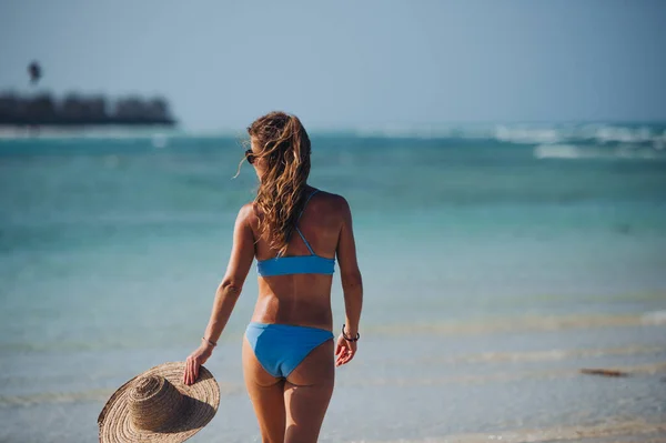 一个身穿比基尼 头戴草帽 身材俊俏的女人在海滩上散步的后视镜 身着蓝色泳衣的女子拍摄了四分之三的照片 她喜欢沙滩和水晶般的大海 — 图库照片