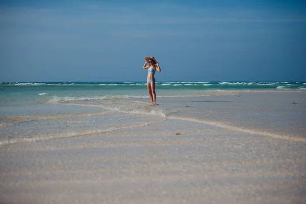 一个穿着比基尼和草帽站在海滩上欣赏阳光的美丽瘦弱女人的画像 全身上下都是身穿蓝色泳衣 戴着太阳镜 享受沙滩和水晶大海的女人 — 图库照片