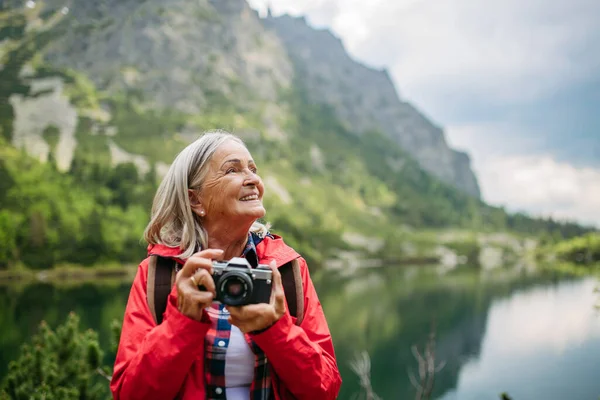 秋の山でハイキングするアクティブなシニア女性の肖像 シニアフレンドリーなトレイル アナログカメラで写真を撮るバックパック付きシニアツーリスト — ストック写真