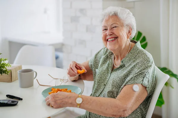 連続的なグルコースモニターを使用して糖尿病のシニア患者は 自宅で血糖値をチェックします 彼女の血糖値を正常に上げるのを助けるために果物を食べるシニアの女性 高齢女性の生命を作るCgmデバイス — ストック写真