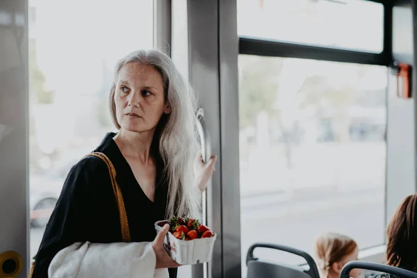一个有着长长的白发的漂亮成熟女人的画像 坐公共汽车旅行 这个成熟的女人站在公共汽车上 手里拿着扶手 提着一盒新鲜草莓 — 图库照片