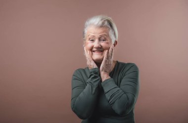 Kısa gri saçlı, yüzünü tutan gülümseyen yaşlı bir kadının stüdyo portresi. Kahverengi pembe arka planda izole edilmiş. Boşluğu kopyala.