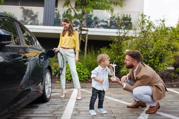 Portriat of woman charging electric car on the street, while husband and son waiting. Happy family in front house waiting to charge electric vehicle. Little boy blowing on wind turbine model. Teaching
