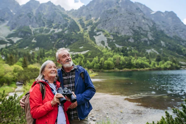 活発なシニアの女性の肖像画は 自然の中で双眼鏡を通して見ています 高齢のカップルは シニアフレンドリーなトレイルで秋の山でハイキング 安定性のためのトレッキングポールを使用してバックパック付きシニアツーリスト — ストック写真
