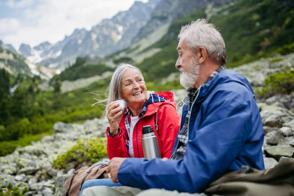 Active elderly couple hiking together in autumn mountains, on senior friendly trail. Husband and wife rehydrating, enjoying warm tea, coffee from thermos. Senior tourist with backpack getting rest