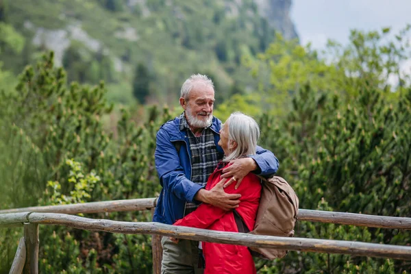 Portrait of beautiful active elderly couple hiking together in autumn mountains, standing on wooden bridge. Senior spouses on the vacation in the mountains celebrating anniversary. Senior tourists are