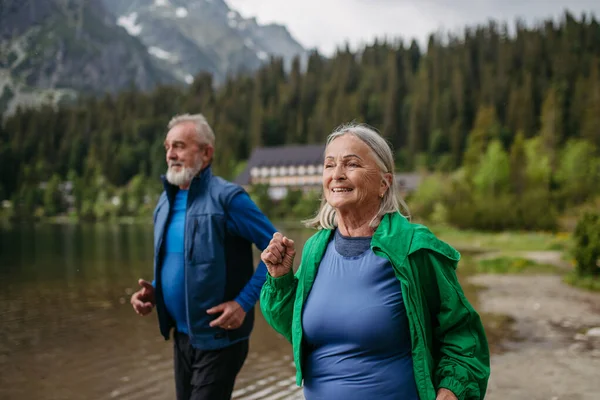 Senior couple running by the lake in the autumn. Elderly husband and wife spending active vacation in the mountains, enjoying combination of physical activity and relaxation.