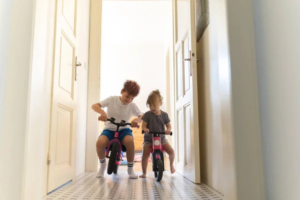 姉妹たちは家の廊下で自転車に乗っている 若い男の子と女の子は自宅で休暇中に楽しんでおり お互いに自転車でレースをしています 兄弟愛と兄弟姉妹の概念 — ストック写真