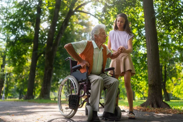 若い介護者は 市の公園で孤独なシニアクライアントと質の高い時間を過ごしています 祖父が車椅子から立ち上がるのを手伝ってくれた孫娘 上の男は立って歩こうとしている — ストック写真