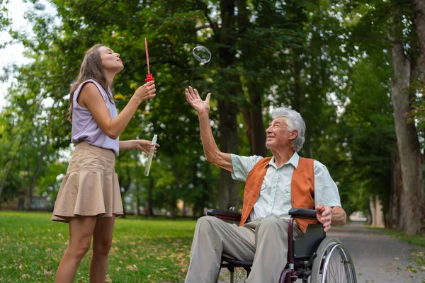 The man in a wheelchair having fun with his caregiver in the park, catching bubbles. The senior man in the wheelchair performs simple exercises, moving as he catches bubbles blown by the young nurse.