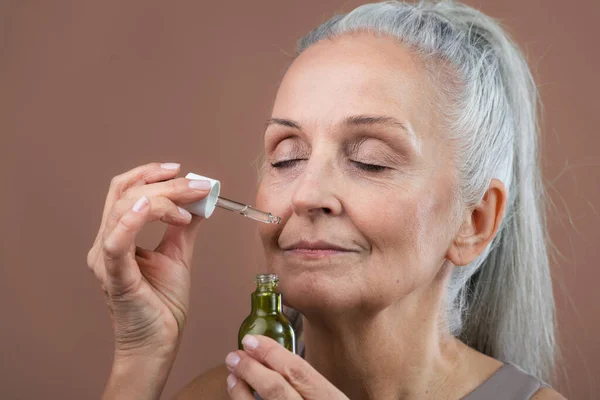 stock image Studio portrait of beautiful senior woman holding skincare serum drops in bottle. Elderly woman taking care of aging skin. Concept of beauty and skincare.