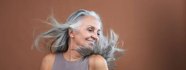 彼女の長い太い灰色の髪を投げる美しいシニアの女性の側面の景色 茶色のピンクのバックグラウンドで孤立 コピースペース付きバナー — ストック写真