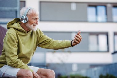 Kıdemli adam şehir dışında egzersiz yapıyor. Yaşlı bir adam bankta dinleniyor, kablosuz kulaklıkla müzik dinliyor, selfie çekiyor..